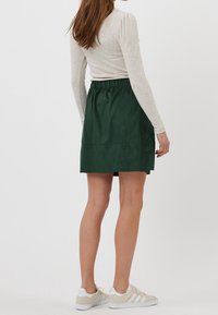 Groene suède rok met een elastische tailleband, kniehoogte en stiksel details. Gecombineerd met beige sneakers en een licht lange mouwen top.