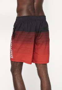 Costumi da bagno con gradiente rosso e nero, dotati di strisce orizzontali, cintura elastica, tasche laterali e un logo riflettente sulla gamba sinistra.