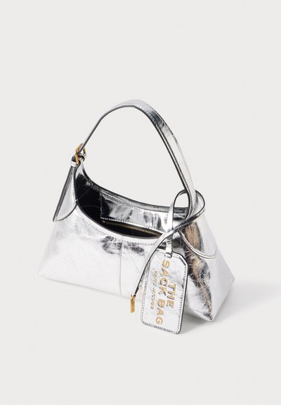 Sac à main argenté brillant avec une poignée unique, une fermeture éclair visible et une étiquette portant la mention "THE SACK BAG MARC JACOBS" en lettres dorées.
