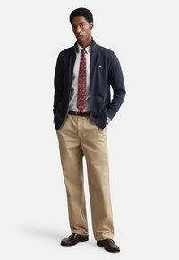 Marineblauw jack met ritssluiting, wit overhemd, rood das met patroon, kaki broek, bruine schoenen. Belangrijke kenmerken zijn de textuur van de stof en de getailleerde pasvorm.