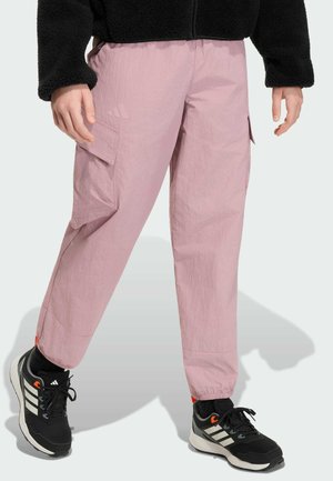 Person trägt hellrosa Cargo-Hose mit elastischen Bündchen, schwarze und weiße Adidas-Sneaker und eine schwarze Fleecejacke.