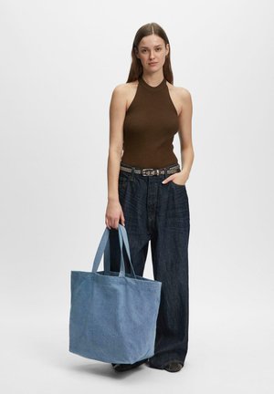 Frau in braunem ärmellosen Oberteil und weiten dunklen Jeans, die eine große blaue Einkaufstasche mit zwei Henkeln vor einfarbigem Hintergrund hält.