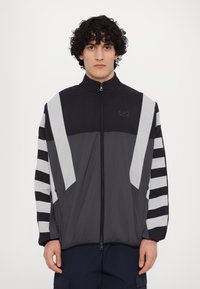 Vindjakke i sort, grå og hvid. Har en høj krave, lynlås lukning og vandrette striber på ærmerne. Letvægts, sporty design.