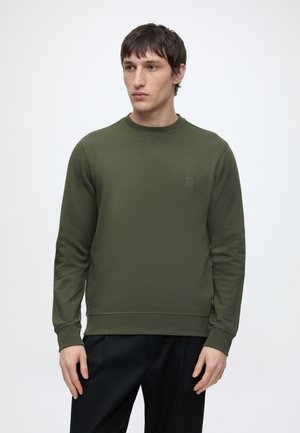 Mladý muž v olivovo zelenom pulóvri s okrúhlym výstrihom a čiernych nohaviciach, stojaci pred jednofarebným svetlosivým pozadím.