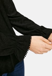 Blusa negra con mangas largas fruncidas que presentan un patrón texturizado y un ribete de pompones en los puños. La tela parece suave y ligera.