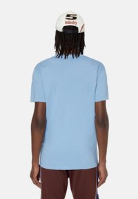 Jasnoniebieska bawełniana koszulka z krótkim rękawem, okrągłym dekoltem, bezszwowa, noszona z brązowymi spodniami i białą czapką z czerwonym napisem.