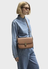 Brun skinnväska med axelrem och klaffstängning, kontrasterande sömmar och guldfärgade beslag; bärs över en jeansoutfit med en skjorta med knappar.