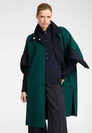 Cappotto in lana verde e navy con colletto alto, caratterizzato da maniche ampie e bottoni decorativi. Indossato sopra un maglione navy e pantaloni a fantasia.