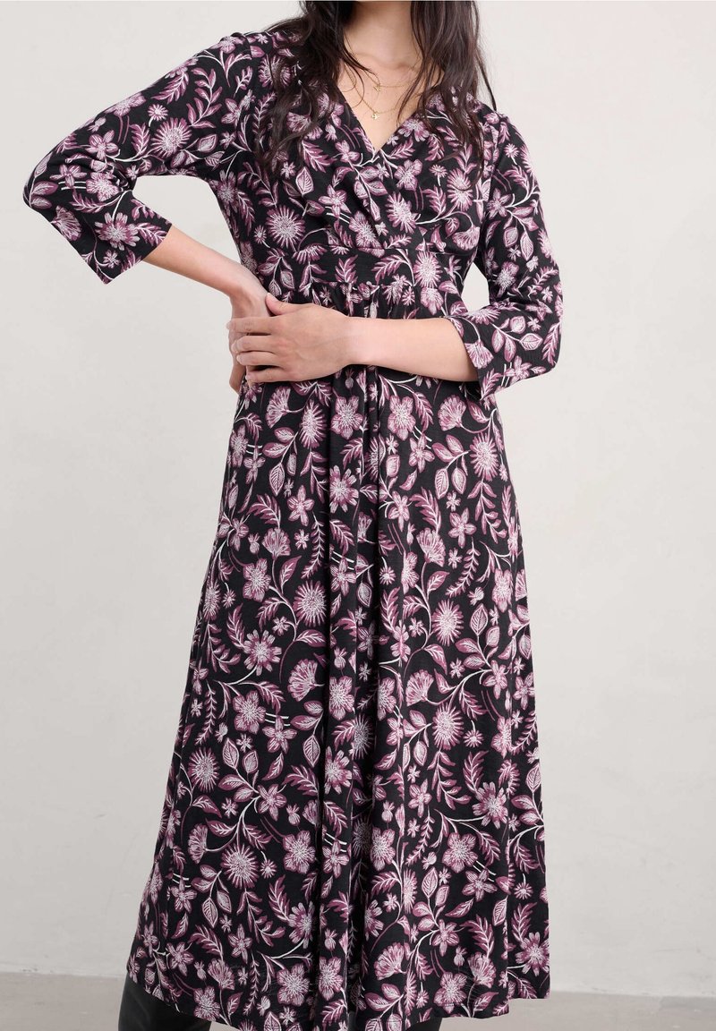 Femme portant une longue robe noire à imprimé floral rose et blanc, debout avec une main sur la taille et l'autre reposant sur son bras.