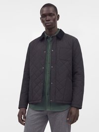 Jeune homme portant une veste matelassée noire par-dessus une chemise verte boutonnée et un pantalon gris, debout devant un fond blanc uni.