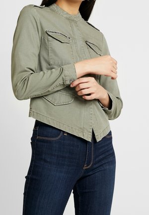 Vrouw draagt een olijfgroene jas met klepzakken en zilveren versieringen, ze past de manchet aan, gecombineerd met donkerblauwe denim jeans.