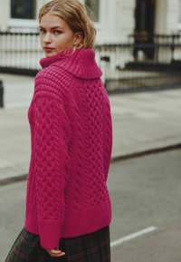 Un pull en tricot câble rose vif avec col haut, motifs texturés et coupe décontractée, associé à une jupe à carreaux sombre.