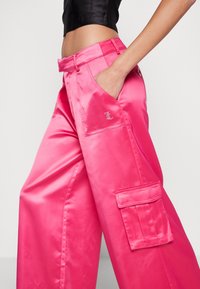 Rosa satin cargo-byxor med avslappnad passform, sidofickor och en dekorativ logga på fickan. Slät textur med en glänsande finish.