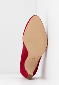 Scarpa con tacco alto in suede rosso, punta affusolata e suola in pelle beige, caratterizzata da una leggera finitura texturizzata e un sottile dettaglio di branding.