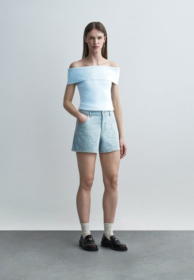 Γυναίκα με λευκό τοπ χωρίς ώμους, ανοιχτό μπλε σορτς ψηλής μέσης με διακοσμητικά, λευκές κάλτσες και μαύρα μοκασίνια στέκεται μπροστά σε απλό φόντο.