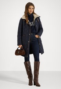 Mornarska kaput s ukrasnim kvadratnim uzorkom i bež umjetnim krznom, uparen s tamnim trapericama i smešnim kožnim čizmama do koljena, drži smeđi suede torbu.