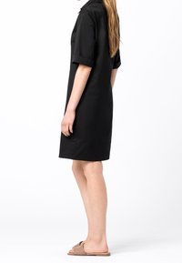 Vestito nero a maniche corte con collo, dalla vestibilità rilassata, caratterizzato da un design semplice e un orlo all'altezza del ginocchio. La modella lo abbina a sandali marroni senza lacci.