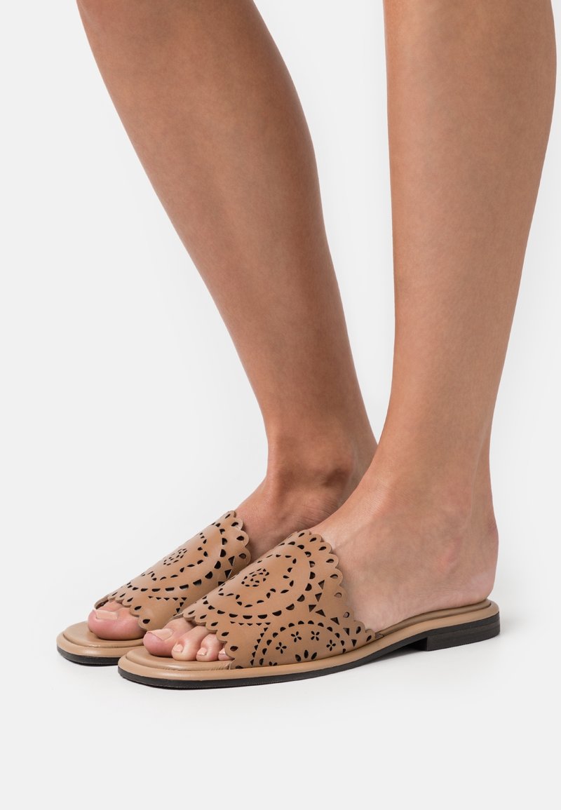 Pieds portant des mules en cuir beige à bouts ouverts avec des motifs floraux découpés au laser et des bords festonnés, semelles plates noires sur fond blanc.