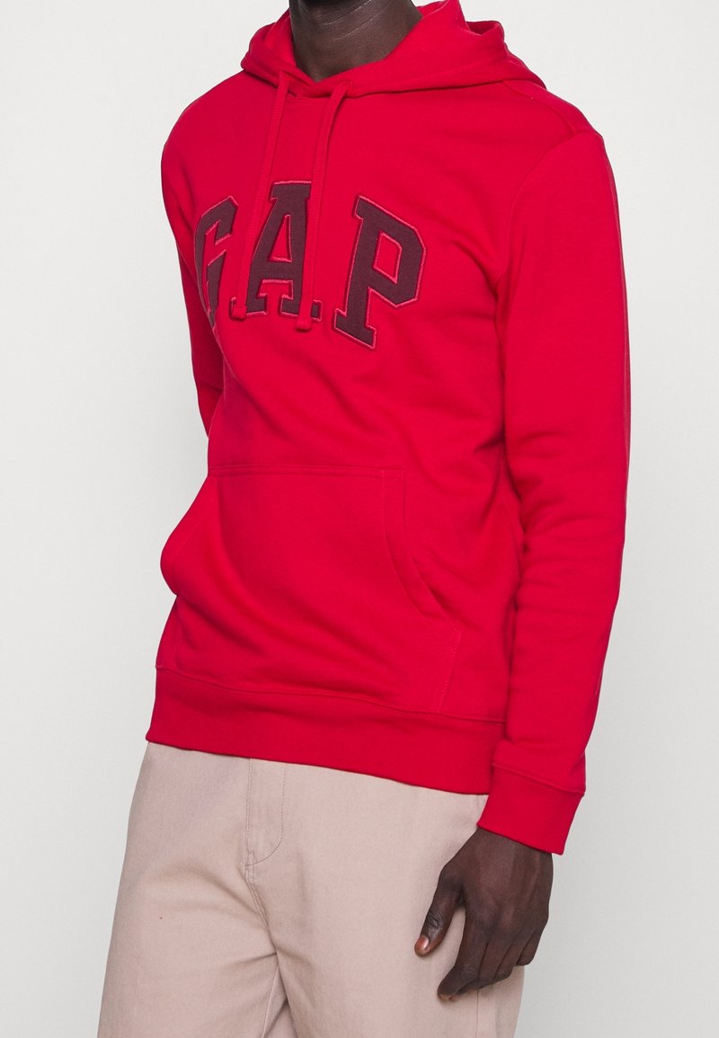 Homme portant un sweat à capuche rouge GAP avec une poche avant et un pantalon beige sur un fond blanc uni.
