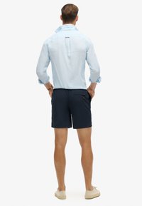 Camisa de lino azul claro con mangas enrolladas, combinada con pantalones cortos oscuros de color marino que tienen un bolsillo trasero. Diseño simple y casual, en colores sólidos.
