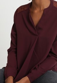 Blusa bordeaux in tessuto leggero con scollo a V e dettagli in pizzo sulle spalle; texture liscia, maniche lunghe, vestibilità comoda.