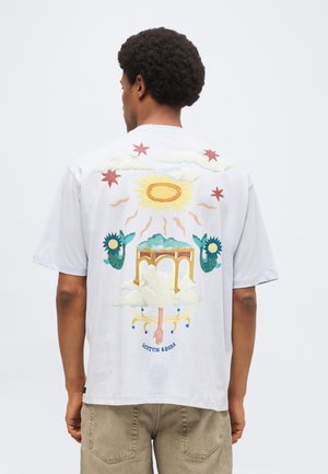 Homme portant un t-shirt blanc avec un graphisme surréaliste coloré comprenant un soleil, des nuages, des arches, des queues de sirènes et une main dans le dos, étiqueté « Scotch & Soda ».