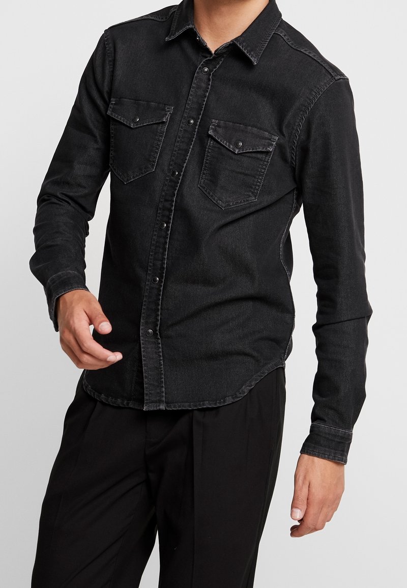 Camicia in denim nera con chiusura a bottoni, due tasche sul petto e maniche lunghe. Presenta un orlo leggermente arcuato e una consistenza morbida.