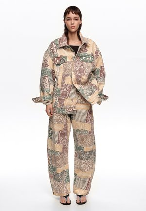 Mujer joven con chaqueta de patchwork oversize a juego y pantalones anchos en tonos verdes apagados, beige y marrón, posando frente a un fondo liso.