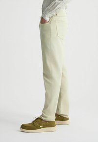 Pantalones color beige claro con corte recto, bolsillos traseros y detalles de costura sutiles, combinados con calzado de ante marrón y una camisa de estampado.