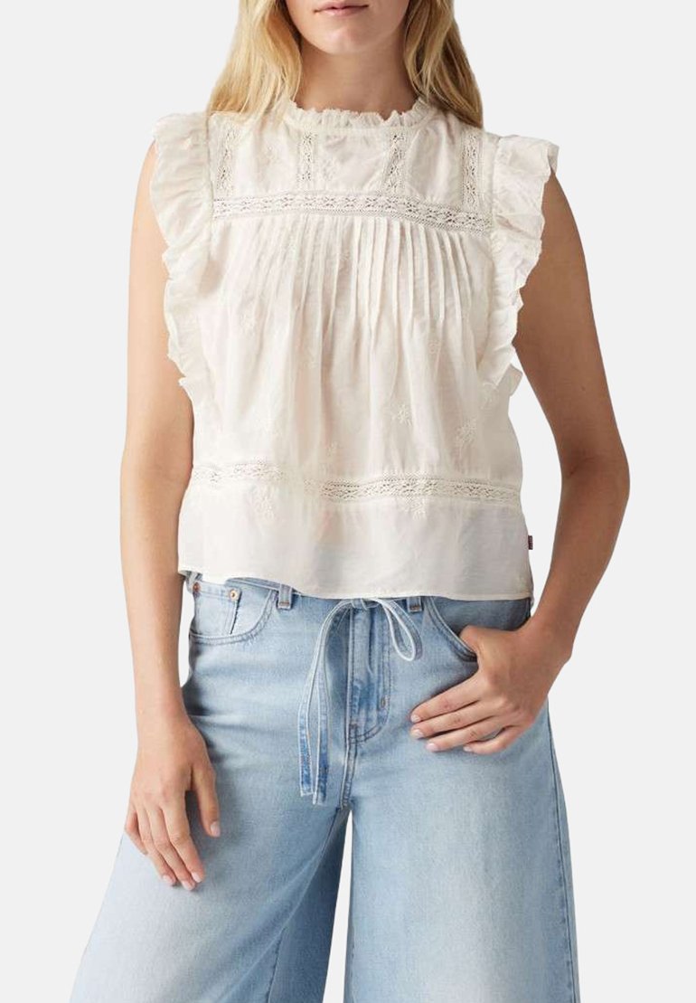 Blouse en coton blanc avec des manches volantes, un devant plissé, des détails en dentelle et une coupe ample, associée à un jean taille haute bleu clair.