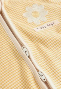 Geltonos ir baltos ginghomo audinys su baltu užtrauktuku, puoštas baltos megztos ramunės motyvu ir etikete su užrašu „laimingos dienos“.