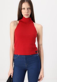 Rdeča pletena brezrokavnica s turtleneck ovratnikom, prilagojena silhueta, rebrast vzorec in oznaka na robu, kombinirana z modrimi kavbojkami.