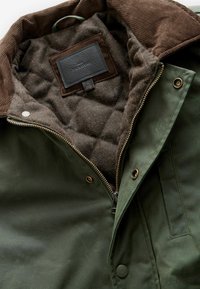 Grüne Outdoor-Jacke mit einem braunen Cordkragen, karierter, wattierter Fütterung und einem Lederetikett; ausgestattet mit einem Reißverschluss und Druckknopfverschlüssen.