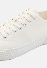 White canvas sneaker with round toe, textured upper, and tonal laces. Features white rubber sole with subtle grooves and metal eyelets.
