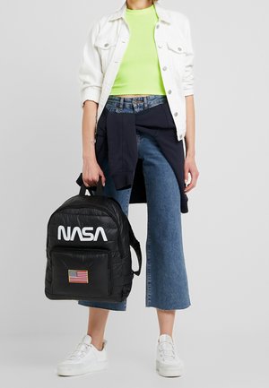 Person holder en sort NASA-rygsæk med patch af det amerikanske flag, iført hvid jakke, neongrøn crop top, blå jeans og hvide sneakers.