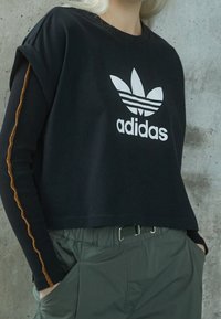 Person wearing black Adidas cropped sweatshirt over long-sleeve shirt with orange seam and green belted pants, standing against concrete wall.