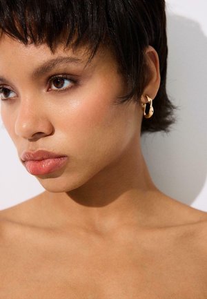 Nuori nainen, jolla on lyhyet tummat hiukset ja lämmin ihon sävy, hänellä on kultaiset renkaankorvakorut ja hän katsoo hieman vasemmalle neutraali ilme kasvoillaan.