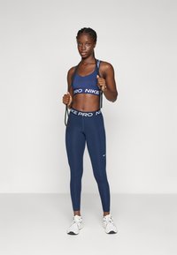 Idrottande kvinna i marinblå Nike sporttop och leggings som håller i ett hopprep, stående på en neutral ljus bakgrund med vita sneakers.
