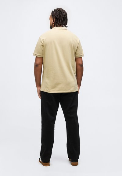 Άνδρας με πλεξούδες μαλλιών που στέκεται με την πλάτη γυρισμένη, φοράει μπεζ κοντομάνικο πόλο πουκάμισο, μαύρο παντελόνι και μαύρα αθλητικά παπούτσια με καφέ σόλες.