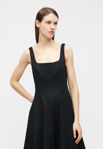 Γυναίκα με ίσια καστανά μαλλιά που φοράει μαύρο αμάνικο φόρεμα με τετράγωνη λαιμόκοψη, στέκεται μπροστά σε απλό φωτεινό φόντο.