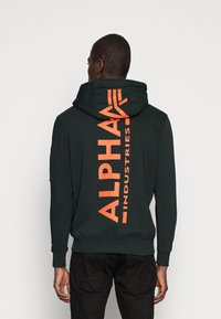 Tamnozelena kapuljača s narančastim tekstom "ALPHA INDUSTRIES" na leđima, sa kenguruh džepom i rebrastim manžetama. Glatka tekstura.