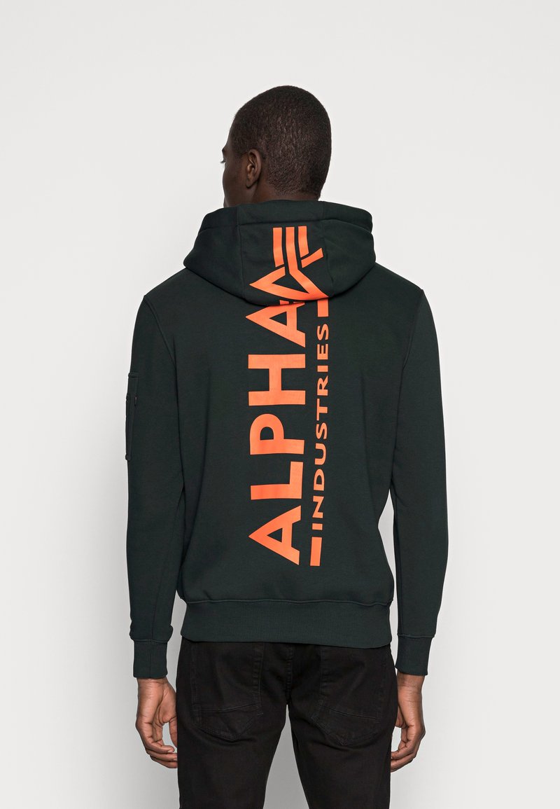Tamnozelena kapuljača s narančastim tekstom "ALPHA INDUSTRIES" na leđima, sa kenguruh džepom i rebrastim manžetama. Glatka tekstura.