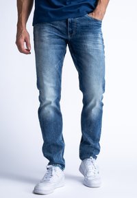 Jasnoniebieskie dżinsy z wyblakłymi miejscami, dopasowane, w pięć kieszeni, z przeszyciami i standardowym stanem. Noszone z białymi sneakersami.