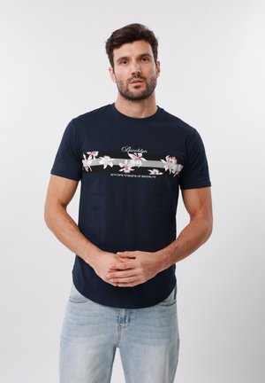 Sötét hajú és szakállas férfi, aki sötétkék pólót visel rózsaszín virágokkal és "Brooklyn" felirattal, fehér egyszínű háttér előtt áll.