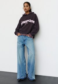 Tummanharmaa huppari, jossa on vaaleanpunaiset kirjaimet, päällysvaatteena raidallisen pitkähihainen paidan päällä. Vaaleansiniset leveälahkeiset farkut, jossa repaleiset reunat ja mustat korkokengät.