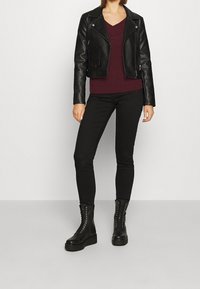 Femme portant une veste en cuir noire, un pull bordeaux à col en V, un pantalon skinny noir et des bottines à lacets noires, debout devant un fond uni.