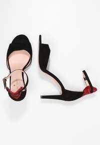 Czarne sandały na wysokim obcasie z paskiem na kostkę, otwartym noskiem i czerwonym brokatowym detalem na pięcie na białym tle.