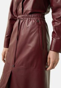 Bordeaux leren rok met een elastische tailleband, zijzakken en een split aan de zijkant. Glad textuur met een subtiele glans en nette stikdetails.