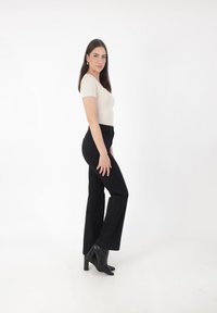 Top beige de manga corta, jeans negros de tiro alto y botas de tacón negras. La modelo está de perfil contra un fondo blanco.