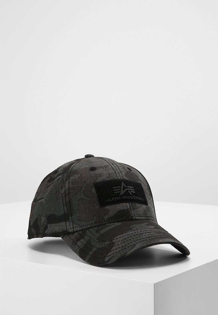 Casquette de baseball camouflage foncé avec patch logo Alpha Industries à l'avant, posée sur une surface blanche sur un fond uni.